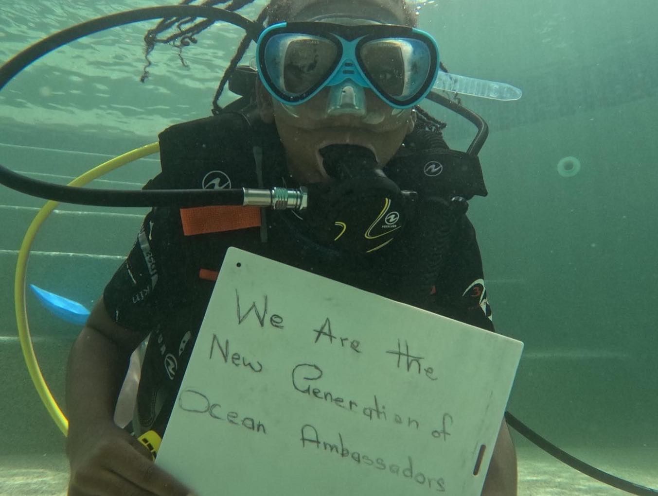 A young diver holds a sign reading We Are the New Generation of Ocean Ambassadors